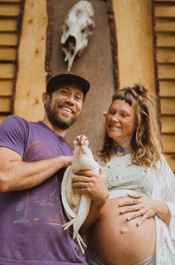 A smiling couple, Robert and Savanna Stark, holding a white chicken in front of a rustic wooden background. Savanna is pregnant, wearing a light dress, while Robert is in a purple t-shirt and a black cap.